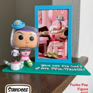 May include: A Funko Pop figure stand featuring a teal base with the text "You see the hat? I am Mrs. Nesbitt!" and a picture frame. The stand holds a Buzz Lightyear Funko Pop figure wearing a blue hat and holding a teacup. The Funko Pop figure is not included.