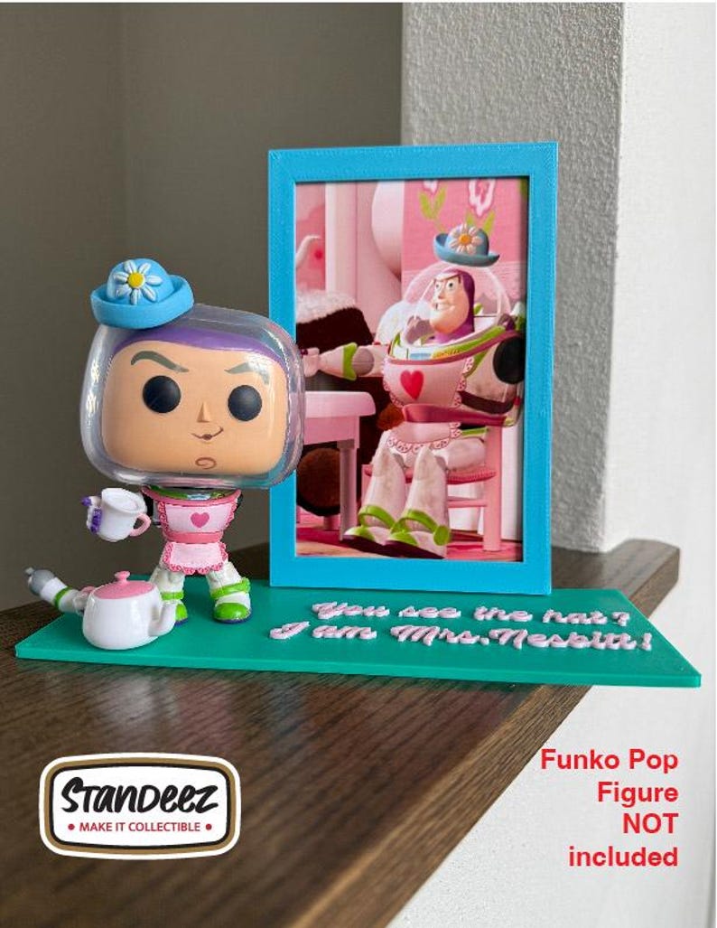 May include: A Funko Pop figure stand featuring a teal base with the text "You see the hat? I am Mrs. Nesbitt!" and a picture frame. The stand holds a Buzz Lightyear Funko Pop figure wearing a blue hat and holding a teacup. The Funko Pop figure is not included.