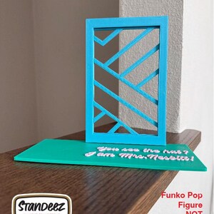 May include: A teal-colored display stand with a geometric design and a teal base. The base has the text "You see the hat? I am Mrs. Nesbitt!" in white. A logo for Standeez is in the lower left corner. The text "Funko Pop Figure NOT included" is in the lower right.
