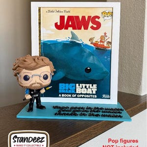 Puede incluir: Un expositor enmarcado con una portada del libro "Jaws" y una figura Funko Pop. La portada muestra un tiburón y un barco. El expositor está sobre una base azul con texto. "Pop figures NOT included."
