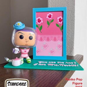 May include: A collectible display stand featuring a Funko Pop figure of Mrs. Nesbit from Toy Story. The stand is teal with a pink and blue framed picture and the text "You see the hat? I am Mrs. Nesbit!"