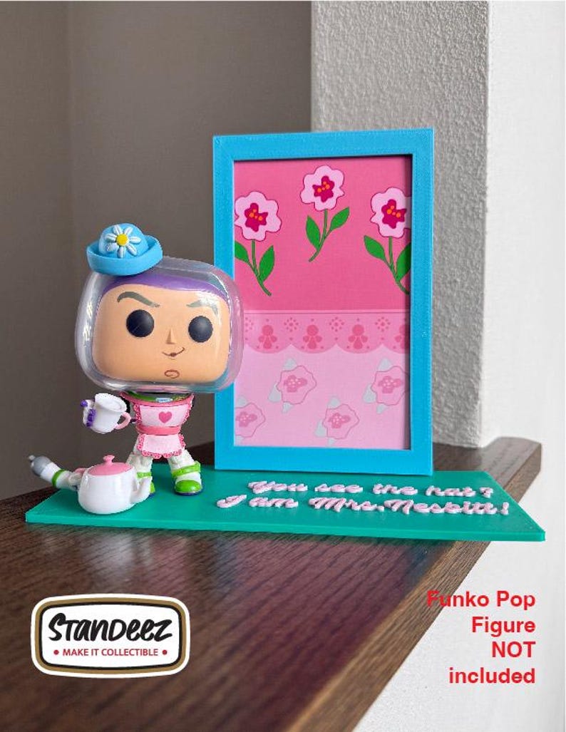 May include: A collectible display stand featuring a Funko Pop figure of Mrs. Nesbit from Toy Story. The stand is teal with a pink and blue framed picture and the text "You see the hat? I am Mrs. Nesbit!"
