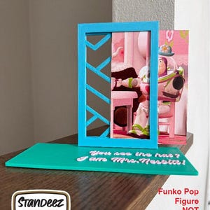 May include: A teal display stand with a blue frame and a printed image of a character. The stand has the text "You see the hat? I am Mrs. Nesbitt!" The Funko Pop figure is not included.