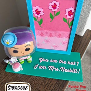 May include: A teal display stand with the text "You see the hat? I am Mrs. Nesbitt!" and a pink floral backdrop. A Funko Pop figure is next to the stand. The stand has a blue frame. The Funko Pop figure is not included.