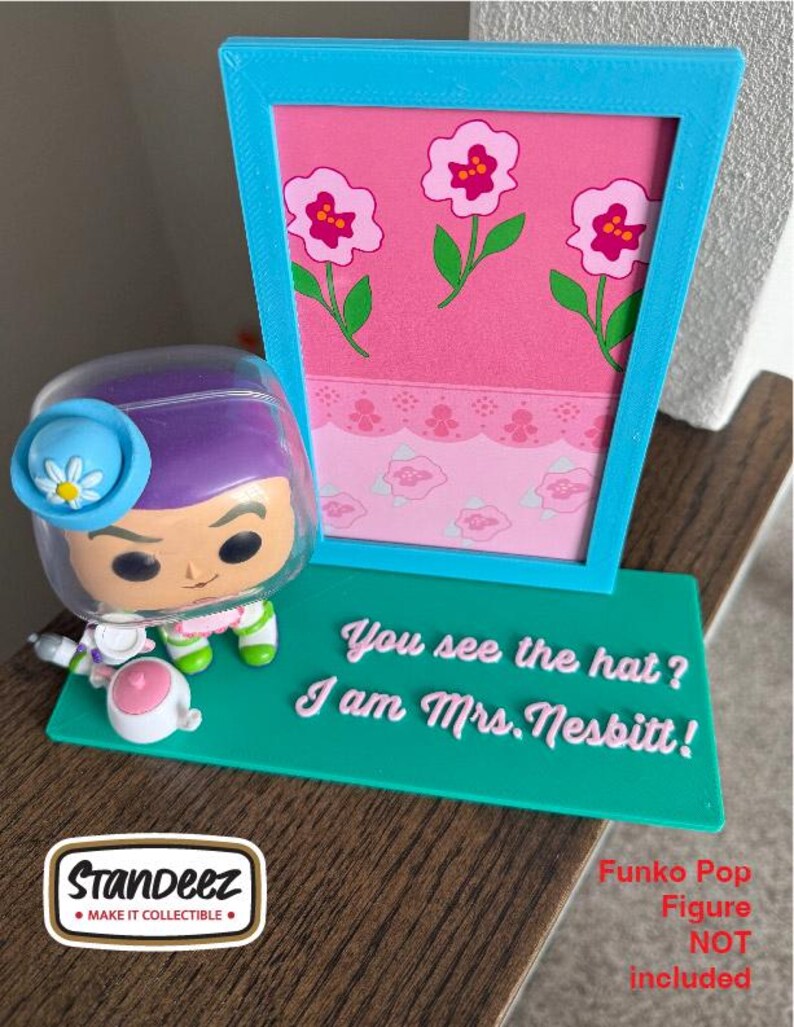 May include: A teal display stand with the text "You see the hat? I am Mrs. Nesbitt!" and a pink floral backdrop. A Funko Pop figure is next to the stand. The stand has a blue frame. The Funko Pop figure is not included.