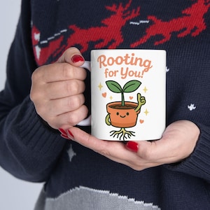 以下が含まれることがあります： 「Rooting for You!」の文字が入った、テラコッタの鉢植えに入った漫画の植物が描かれた白いセラミックマグカップ。植物は緑色の葉を持ち、親指を立てています。マグカップは可愛らしいデザインです。