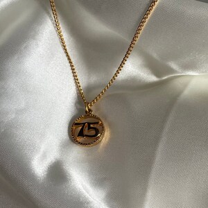 May include: A gold-colored necklace with a delicate chain and a circular pendant. The pendant features the number "75" in a black, stylized font, encircled by a textured border. The necklace rests on a white, silky fabric.