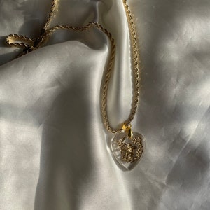 May include: Gold-coloured necklace with a clear heart-shaped pendant. The pendant features a gold design of a bow and arrow within a wreath. The chain has a rope-like texture, and the necklace rests on a white satin surface.