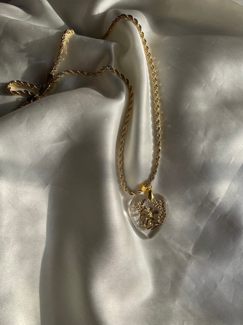May include: Gold-coloured necklace with a clear heart-shaped pendant. The pendant features a gold design of a bow and arrow within a wreath. The chain has a rope-like texture, and the necklace rests on a white satin surface.
