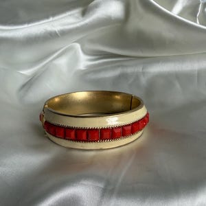 May include: A hinged bangle bracelet featuring a cream-colored enamel finish and a central row of square, bright red stones. The bracelet is accented with thin, gold-toned metal trim along the edges of the red stone inlay and has a gold interior.