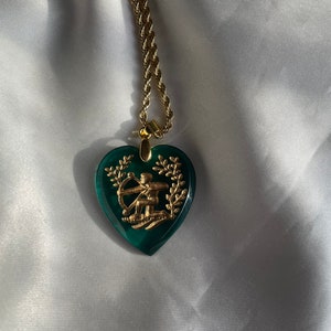 May include: Gold-coloured necklace featuring a heart-shaped pendant. The pendant is a deep green with a gold design of a figure with a bow and arrow, framed by a wreath. The chain is a twisted rope style.