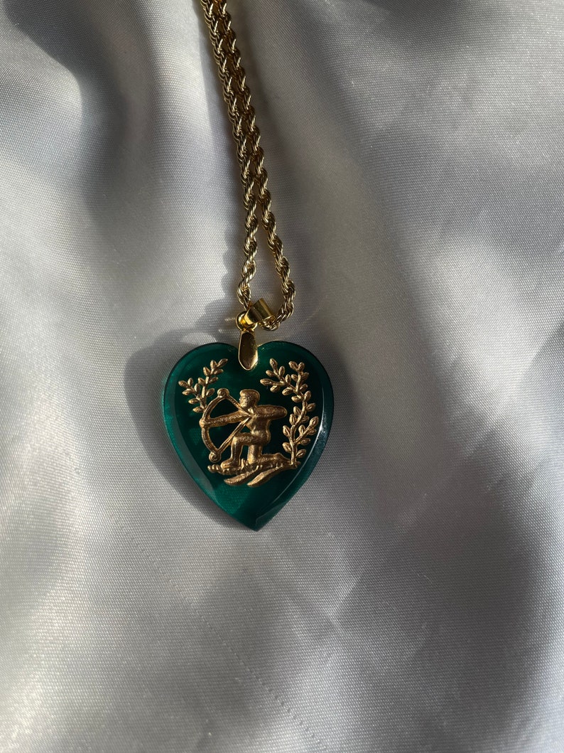 May include: Gold-coloured necklace featuring a heart-shaped pendant. The pendant is a deep green with a gold design of a figure with a bow and arrow, framed by a wreath. The chain is a twisted rope style.