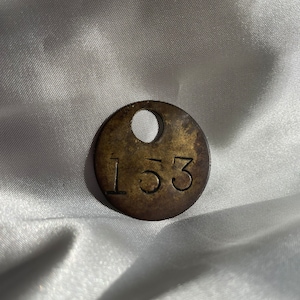 May include: A circular, antique brass tag with the number "153" stamped on it. The tag has a small hole at the top and shows signs of age and wear. The background is a white, textured fabric.