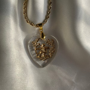 May include: A gold necklace with a clear heart-shaped pendant. The pendant features a gold archer with a bow and arrow, surrounded by leaves. The chain has a twisted rope design.