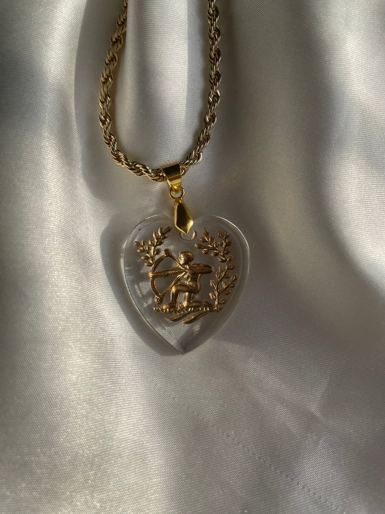 May include: A gold necklace with a clear heart-shaped pendant. The pendant features a gold archer with a bow and arrow, surrounded by leaves. The chain has a twisted rope design.
