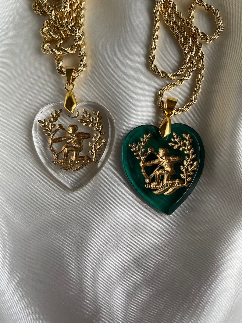May include: Two heart-shaped pendants on gold chains. One clear, one green. Each has a gold archer design within a gold laurel wreath. The chains are gold rope style.