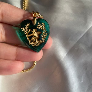 May include: Gold necklace featuring a heart-shaped pendant. The dark green pendant has a gold Sagittarius design and laurel leaves. The chain is a delicate rope style.