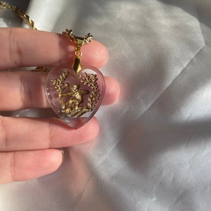May include: A gold-coloured necklace with a heart-shaped pendant. The pendant features a gold-coloured archer figure and laurel wreath design. The necklace is on a gold-coloured chain.