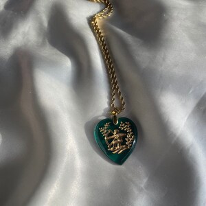 May include: A gold chain necklace featuring a heart-shaped, emerald green pendant. The pendant has a gold-coloured design of a person with a bow and arrow. The necklace is displayed on a white, silky fabric.