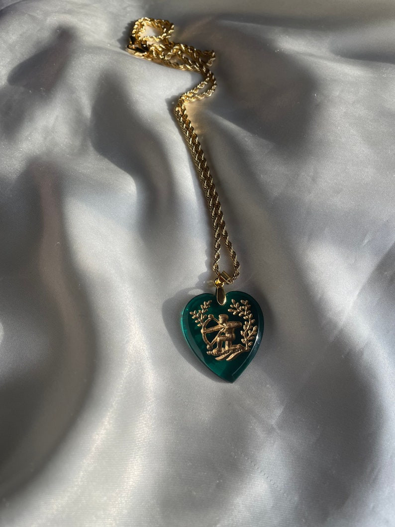 May include: A gold chain necklace featuring a heart-shaped, emerald green pendant. The pendant has a gold-coloured design of a person with a bow and arrow. The necklace is displayed on a white, silky fabric.