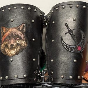 May include: Pair of black leather arm guards with silver rivets. One guard features a fox face, the other a sword through a crescent moon. The guards are likely for cosplay or historical reenactment.
