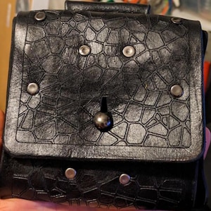 May include: Black leather pouch with a textured, geometric pattern. The pouch has a flap closure secured by a metal ball and is accented with silver-colored rivets. The design suggests a medieval or fantasy theme.