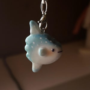 May include: A handcrafted sunfish charm. The charm is light blue with white spots and a white underside. It features a small, black line for a mouth and is attached to a silver-coloured clasp.