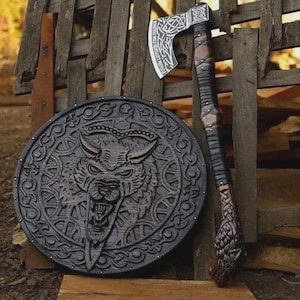 Puede incluir: Un escudo circular de madera con una cabeza de lobo tallada en el centro rodeada de intrincados patrones de nudos, junto a un hacha de mango largo. La cabeza del hacha muestra detalles de metal grabado en tono plateado y un pomo con forma de lobo.