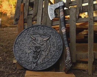 May include: A circular wooden shield featuring a carved wolf head at the center with intricate knotwork patterns, paired with a long-handled axe. The axe head displays detailed metal engravings, while the handle is wrapped in dark leather strips.
