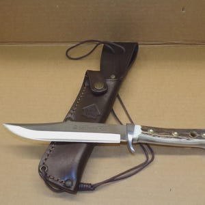 Peut inclure: Un couteau à lame fixe avec une lame en acier argenté et un manche en bois de cerf brun texturé doté de rivets en laiton. Le couteau est placé horizontalement devant un étui en cuir brun foncé avec une fermeture à pression et un cordon en cuir.