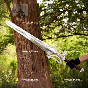 Può includere: Una spada in metallo color argento con lama a doppio taglio e una guardia complessa e curva. L'impugnatura è rivestita in pelle marrone scuro e il pomolo è un semplice tappo in metallo. La spada è tenuta da una mano con un guanto nero.
