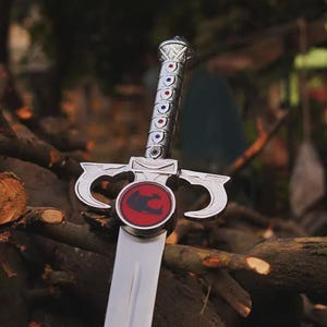 May include: A silver-colored sword featuring a metallic handle with a series of small blue and red circular inlays. The crossguard displays a prominent circular emblem with a black cat-like silhouette centered on a vibrant red background.