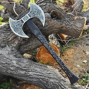 May include: A double-bladed axe with a black handle and silver-colored blades. The blades feature intricate Celtic knot designs. The handle is decorated with small silver-colored floral and star patterns, and a triangular symbol.