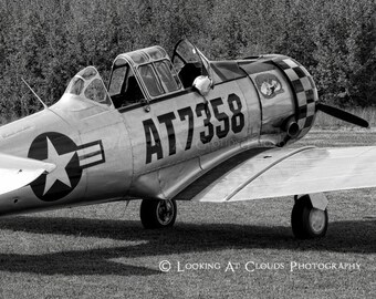 May include: A black and white photo of a vintage silver airplane with the tail number AT7358. The plane has a star and stripes on the side and a checkered pattern on the tail.