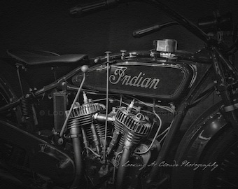 May include: Black and white photo of a vintage Indian motorcycle. The motorcycle has a large, chrome engine and a leather seat. The word "Indian" is written in cursive on the gas tank.