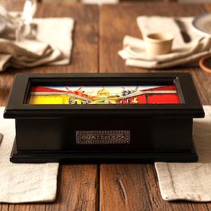 May include: A black wooden box with a colorful image on top, set on a wooden table with place settings. The box has the word "GUATEMALA" on it. Other items include plates, silverware, and a bottle of oil.