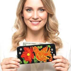 May include: Black wallet with a colorful floral and bird design. The wallet features a red flower and an orange bird, with green accents on a black background. The wallet is held up by a person.