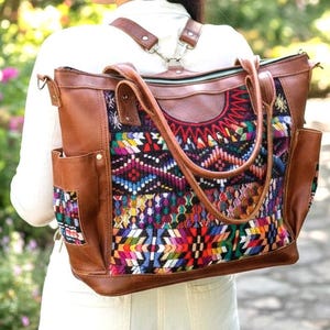 May include: A brown leather backpack with a colourful woven design on the front. The bag has multiple pockets and adjustable straps. The person is wearing a white shirt and walking in a garden.