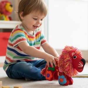 Puede incluir: Un niño pequeño juega con un juguete de peluche de león rojo y naranja. El león tiene una nariz negra y una melena colorida. El niño lleva una camiseta a rayas y vaqueros azules.