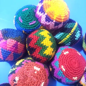 May include: A collection of round, crocheted hacky sacks featuring vibrant, multi-colored geometric patterns. Each ball is crafted with intricate yarn stitching in shades of red, yellow, blue, green, purple, and black, creating a textured surface.
