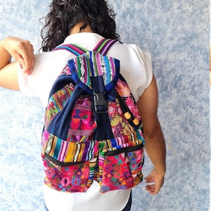 May include: A brightly coloured backpack with a patterned design. The backpack features a mix of colours, including pink, blue, yellow, and green. It has two front pockets with zips, a black buckle closure, and adjustable straps.