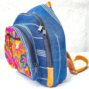 May include: A blue backpack with white stripes and a colorful embroidered design. The backpack has multiple zippered compartments and brown leather accents. The embroidered design features a bird and floral pattern.