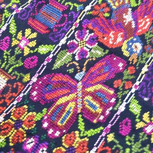 May include: A black fabric featuring a dense, multi-colored cross-stitch embroidery pattern. The design displays vibrant butterflies in shades of purple, red, and yellow, interspersed with various floral motifs in bright green, pink, and orange.