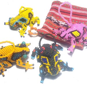 Op de afbeelding: Vier kralen kikker ornamenten in geel, oranje, roze en zwart. De kikkers hebben gedetailleerd kralenwerk met blauwe, zwarte en gele accenten. Een roze kikker is bevestigd aan een rood-witte geweven tas. De kikkers zijn een uniek decoratief item.