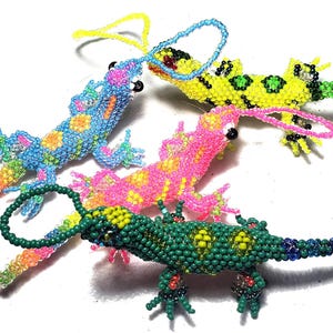 Puede incluir: Cuatro adornos de lagarto con cuentas en colores vibrantes: azul, rosa, amarillo y verde. Cada lagarto tiene un lazo para colgar y está decorado con cuentas de colores. Los lagartos están sobre un fondo blanco.