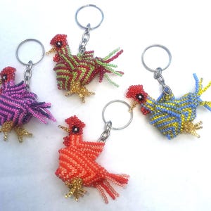 May include: A collection of beaded rooster keychains in various colors. The keychains feature red, green, purple, orange, and blue roosters with metal key rings. The roosters have detailed beadwork and are a decorative accessory.