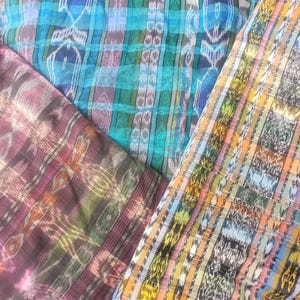 May include: Three pieces of fabric featuring intricate ikat-style woven patterns. One section displays vibrant blue and teal tones, another features deep maroon and earthy brown hues, and the third showcases a multi-coloured plaid with yellow and orange.