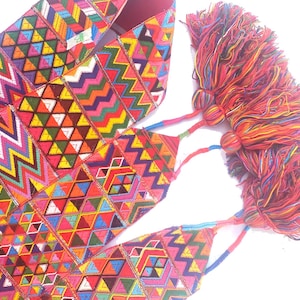 May include: A colorful textile with geometric patterns in red, pink, yellow, blue, and green. The design features zigzags, triangles, and diamond shapes. The textile is adorned with large, vibrant tassels in shades of red, orange, and yellow.