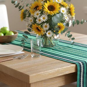 May include: A wooden dining table set for a meal. A green and white striped table runner is centered on the table. A white plate, silverware, and a white napkin with fringe are on the left. A vase of sunflowers and daisies is in the center.