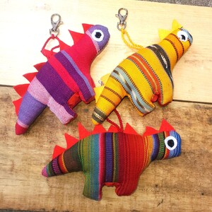 May include: Three colorful dinosaur keychains. Each plush dinosaur has a striped pattern in various colors, red spines, and a metal clasp. The dinosaurs are on a wooden surface.